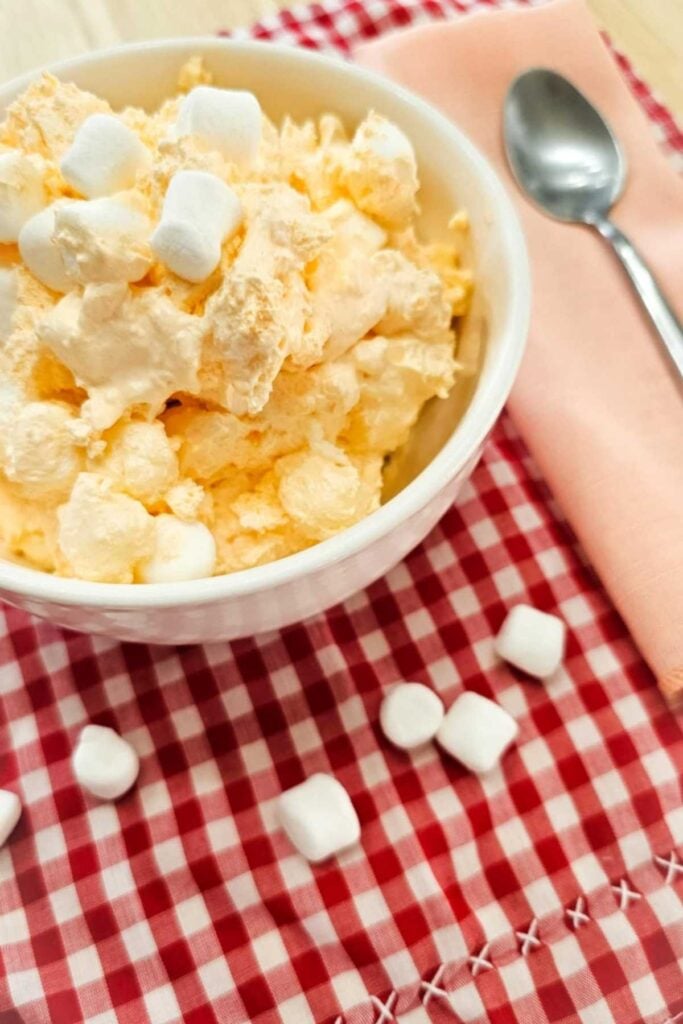 grandmas orange fluff in a white glass bowl topped with marshmallows and on a red and white vintage checkered table cloth with a peach cloth napkin and a spoon