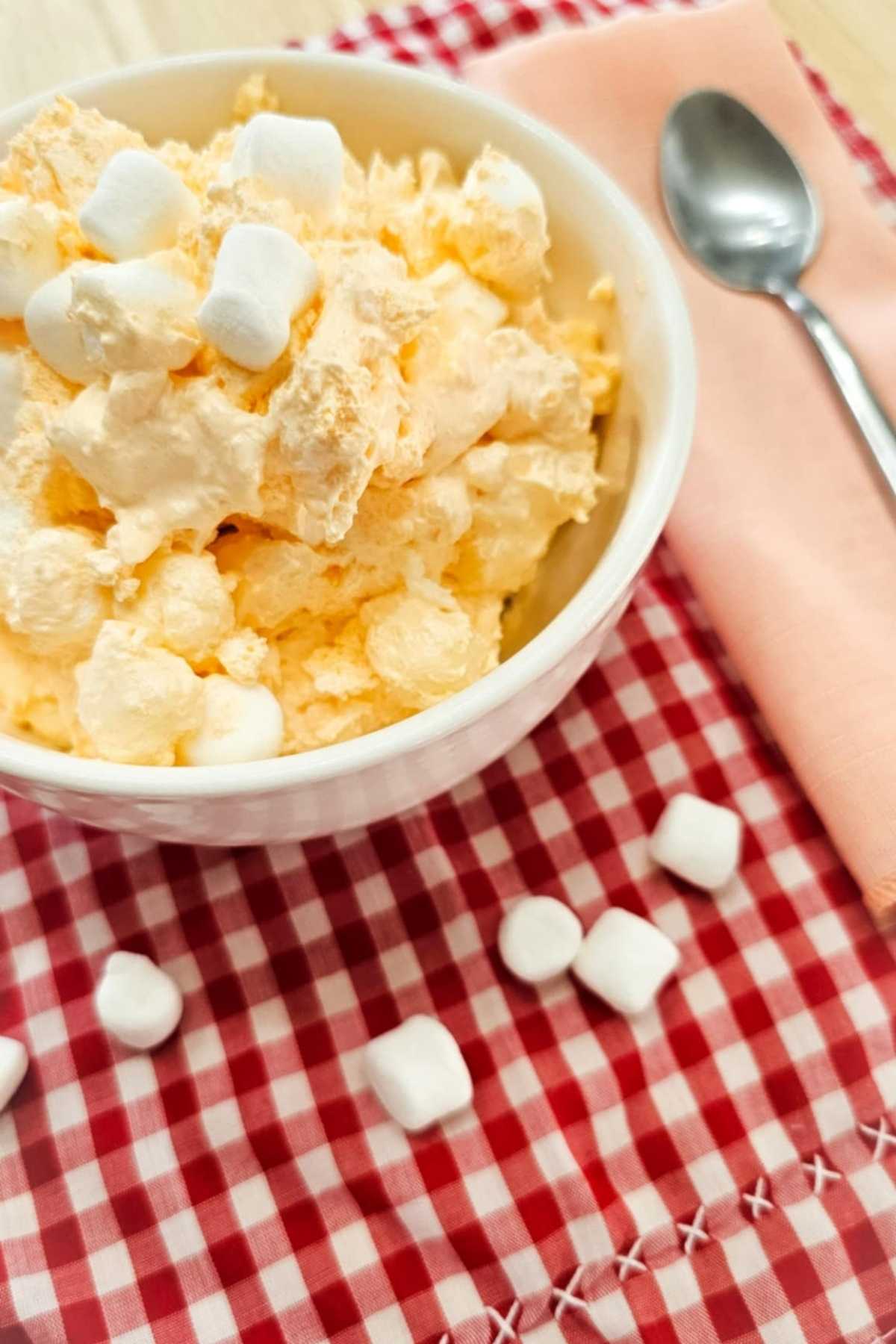 grandmas orange fluff in a white glass bowl topped with marshmallows and on a red and white vintage checkered table cloth with a peach cloth napkin and a spoon