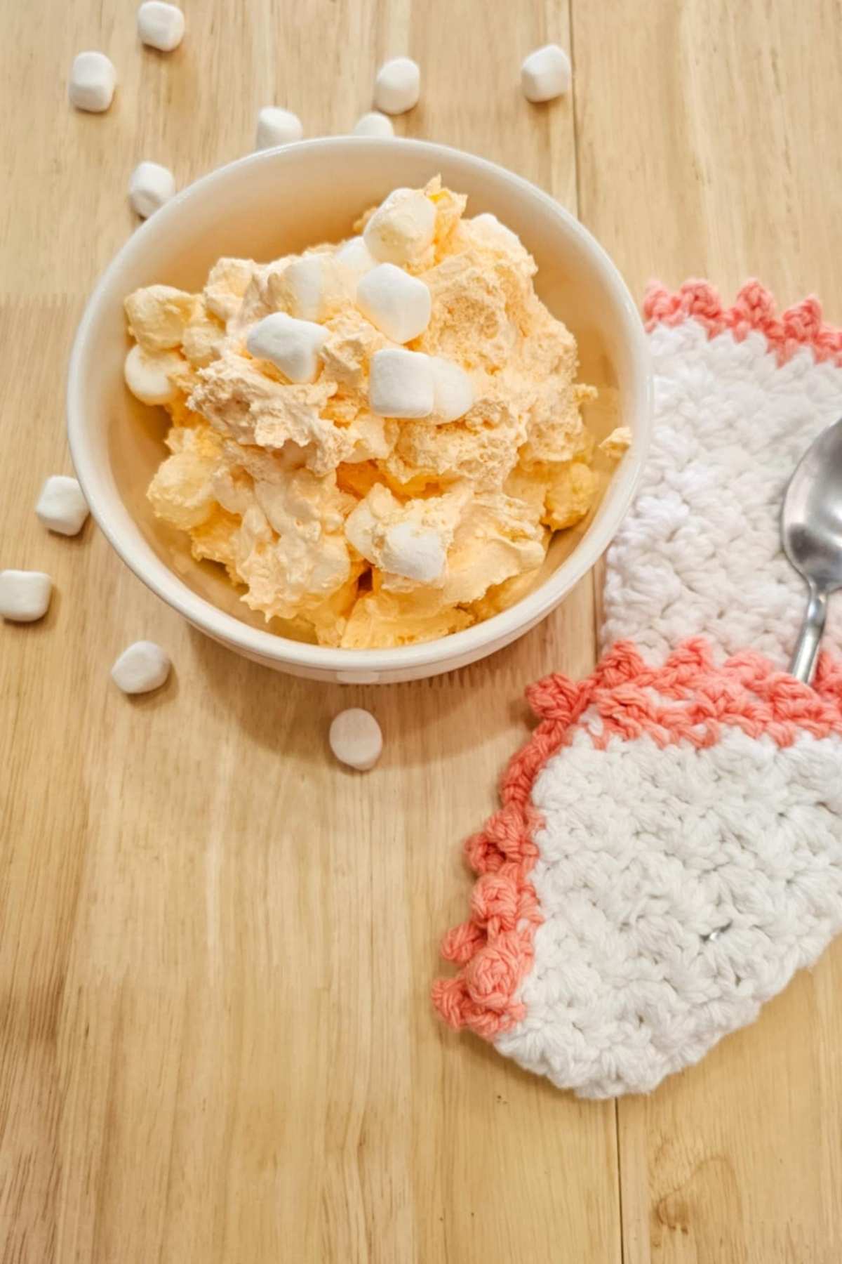 grandmas orange cool whip dessert in a white bowl on a wooden counter top with a white and peach crochet hot pad with a spoon on it