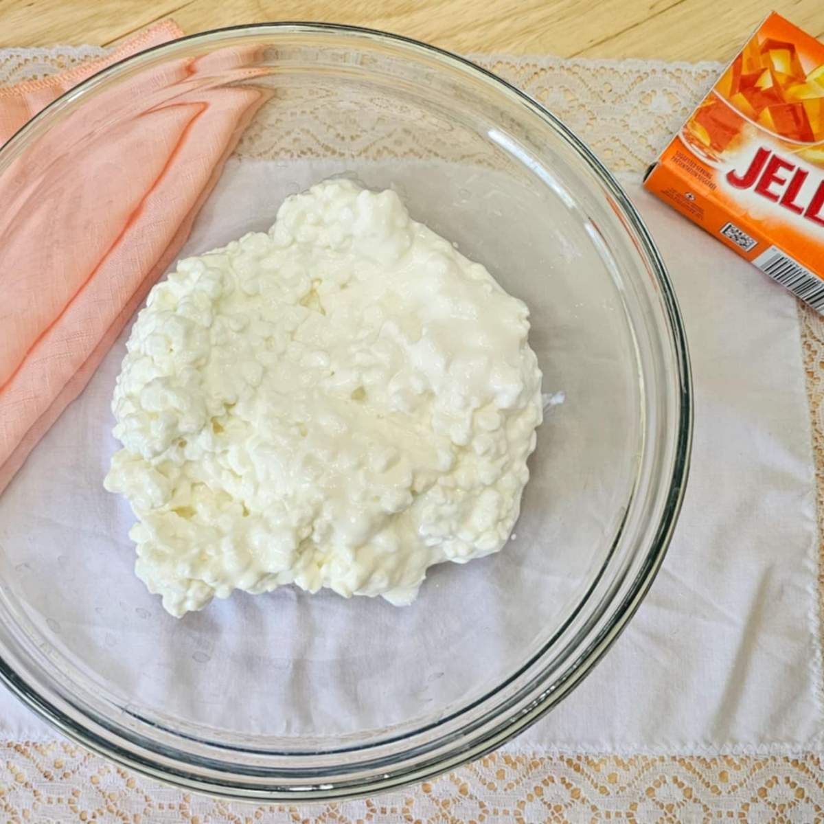 cottage cheese in a clear glass bowl on a vintage white placemat with lace with an orange jello package next to it and a peach cloth napkin
