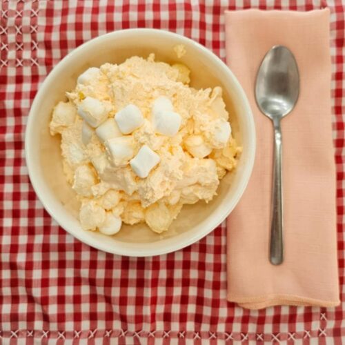 grandmas orange fluff dessert in a white glass bowl, topped with marshmallows on a red and white vintage table cloth with a peach cloth napkin next to it with a stainless steel spoon