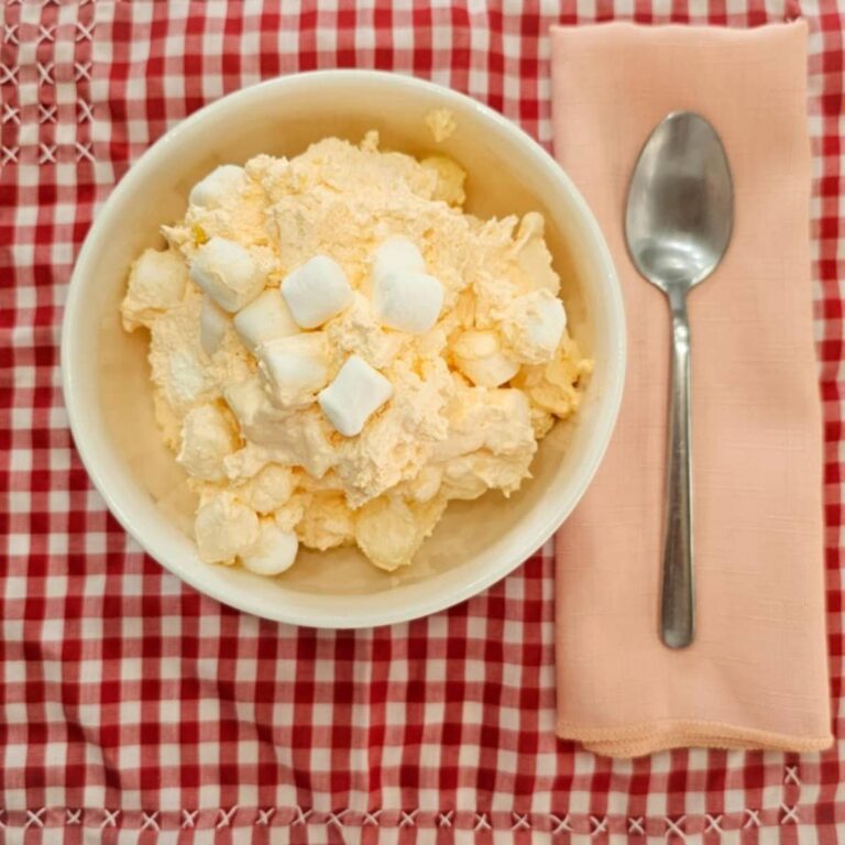 grandmas orange fluff dessert in a white glass bowl, topped with marshmallows on a red and white vintage table cloth with a peach cloth napkin next to it with a stainless steel spoon