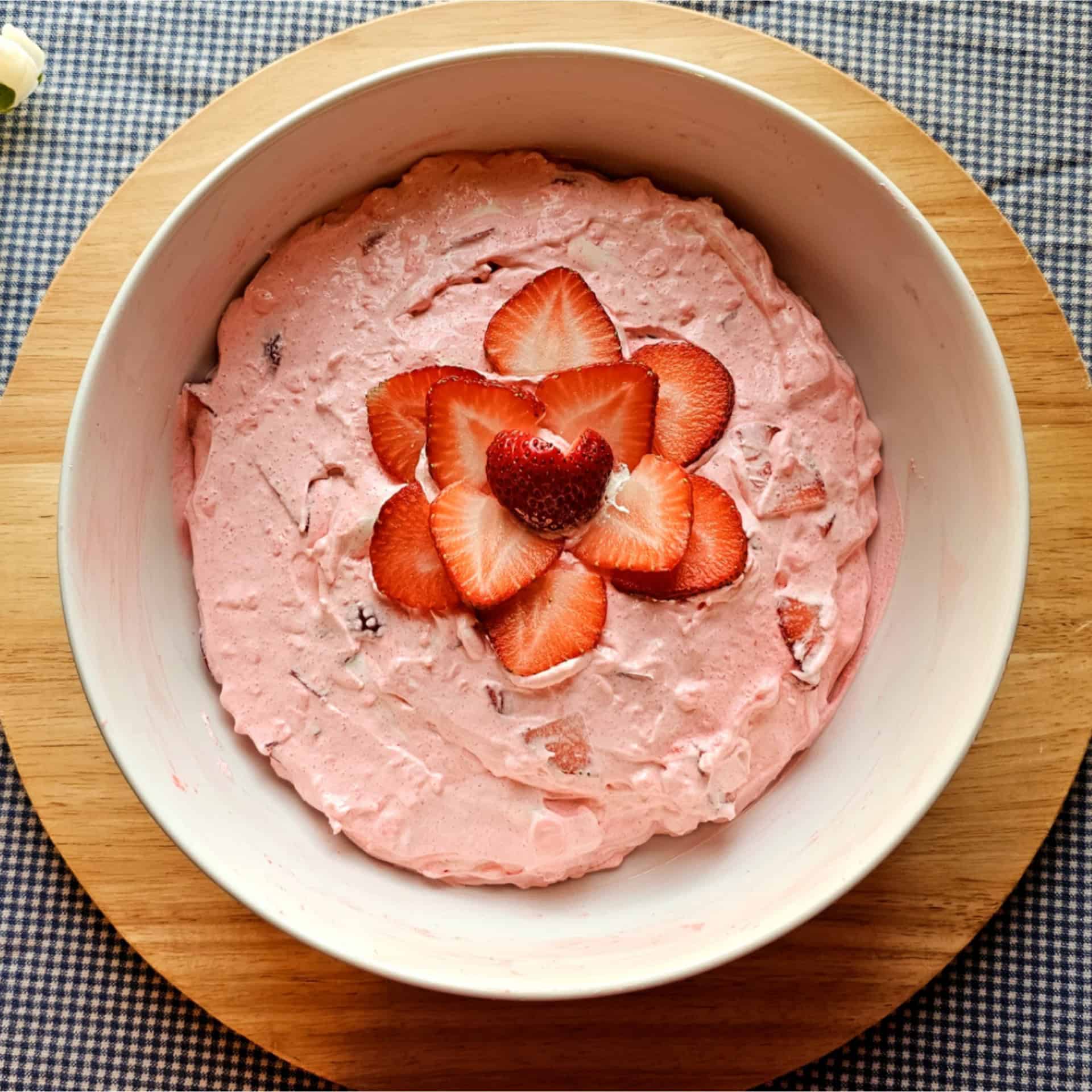 strawberry jello fluff in a white glass bowl garnished with fresh strawberries on a wooden cutting board on a white and blue checkered table cloth