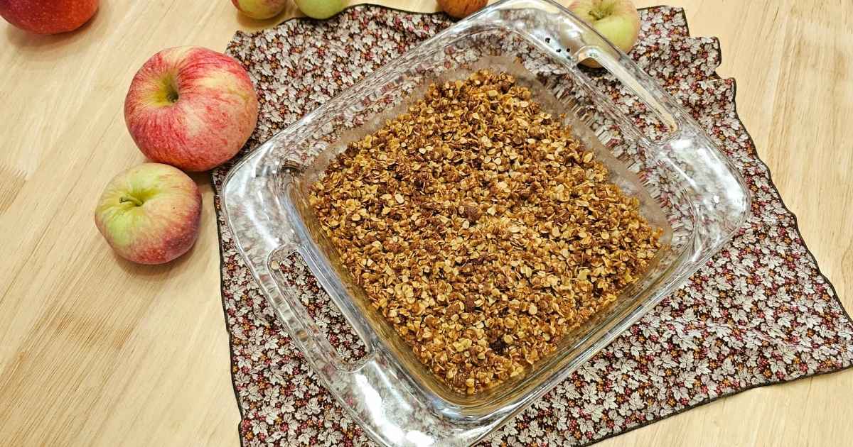 apple crisp in a glass 8x8 dish on a wooden counter top with freshly picked apples to the side of it