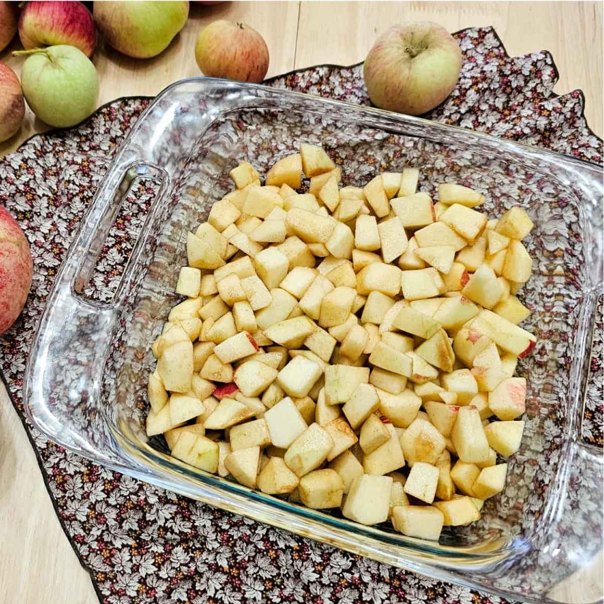 bottom layer of the apple crisp in an 8x8 glass dish sitting on a fall cloth napkin on a wooden counter top with fresh apples to the side of it