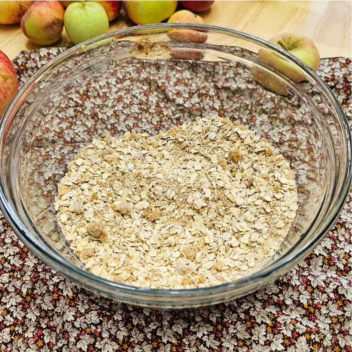 oatmeal, flour and spices in a clear glass bowl on a wooden counter top with apples to the side of it