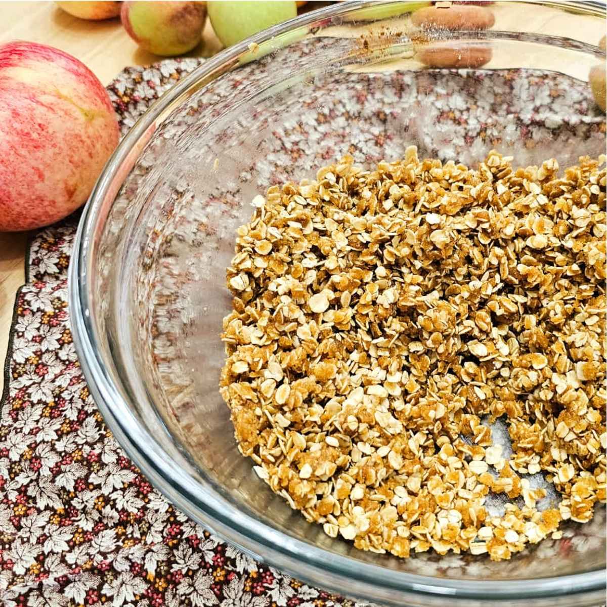 Apple crisp topping in a clear glass bowl on a wooden counter top sitting on a fall cloth napkin with fresh apples to the side of it