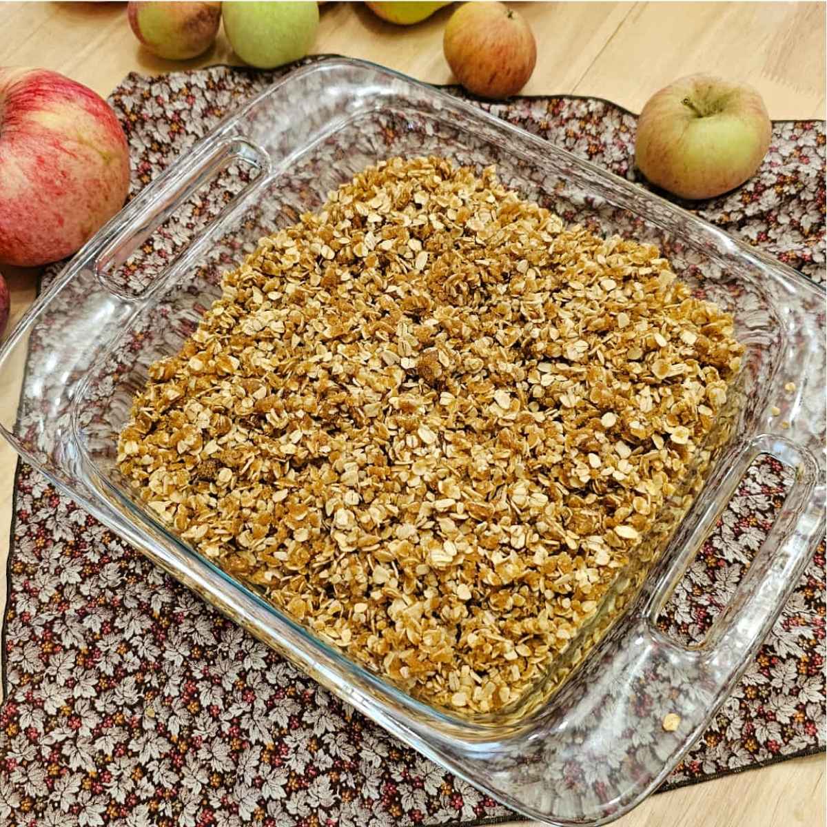apple crisp topping added on top of the bottom layer of apples sitting in an 8x8 glass dish on a wooden counter top with apples to the side of it