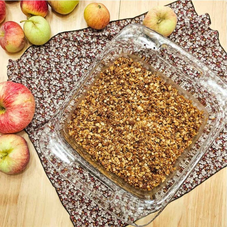 grandmas apple crisp fresh from the oven sitting on a fall cloth napkin with fresh apples to the side of it on a wooden counter top
