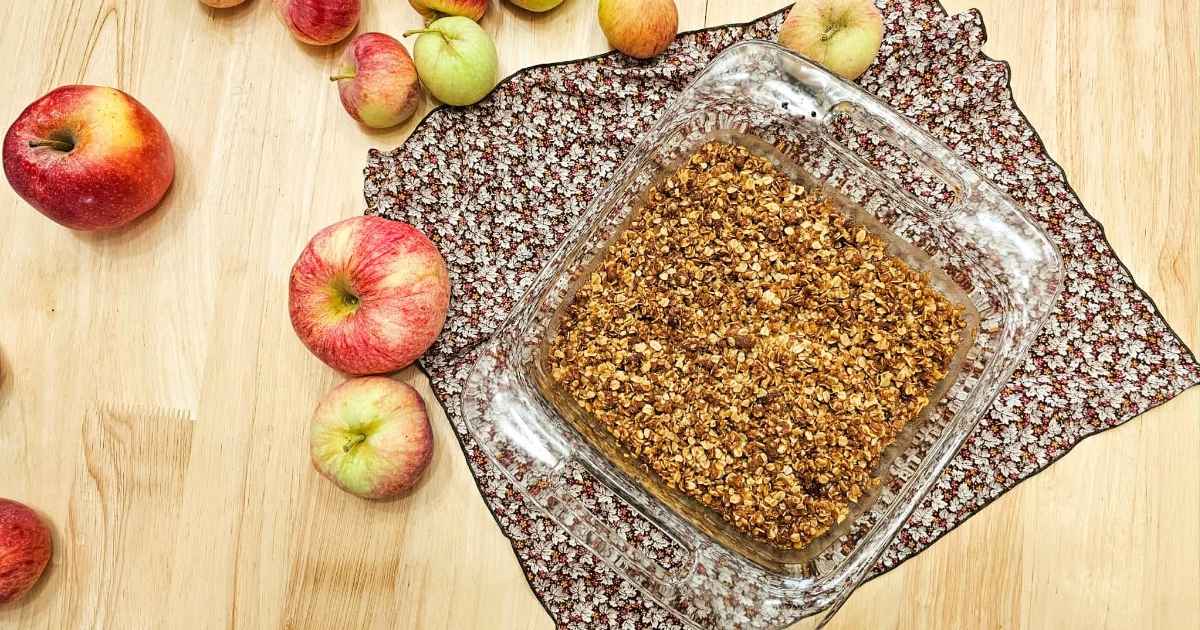 grandmas apple crisp in an 8x8 pan sitting on a fall cloth napkin on a wooden counter and surrounded by freshly picked apples