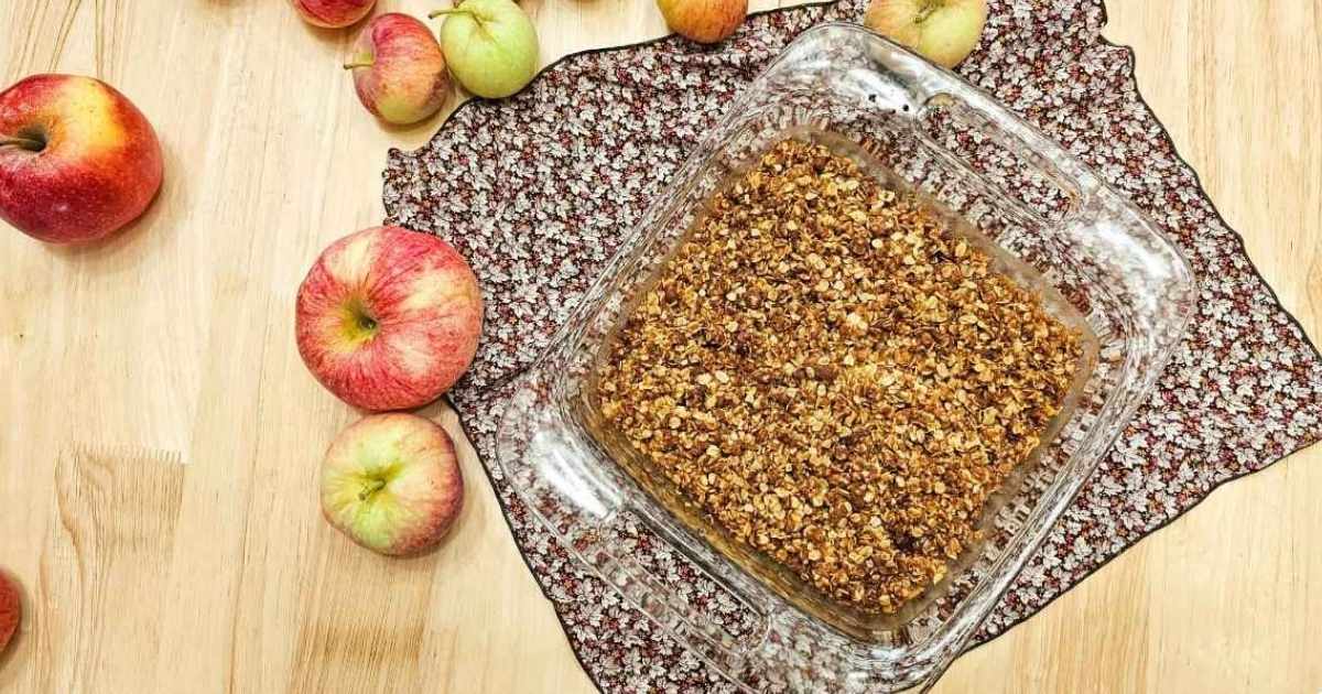 grandmas old fashioned apple crisp in an 8x8 dish on a wooden counter top with apples around it that were freshly picked