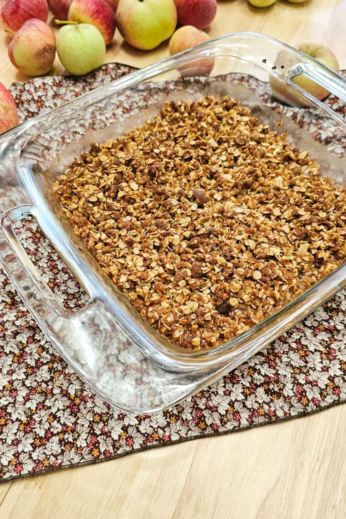 apple crisp in an 8x8 glass dish sitting on a fall coloured cloth napkin with leaves on a wooden counter with fresh apples to the side of it