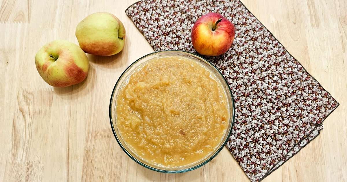 apple sauce in a large clear bowl with fresh apples to the side of it with a fall cloth napkin underneath with leaves on it 