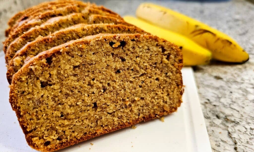 old fashioned banana bread sitting on a white plastic cutting board with bananas in the back ground