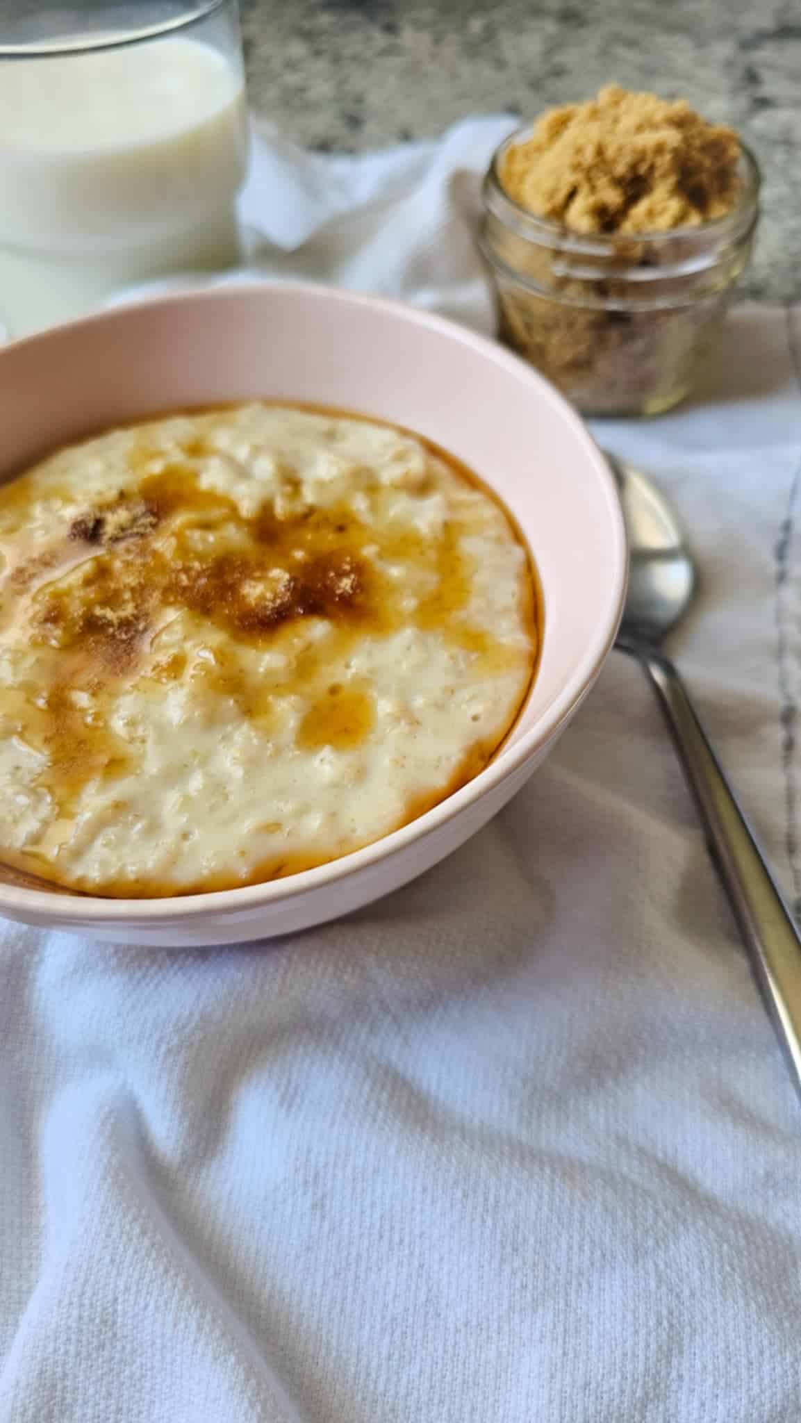 Creamy oatmeal in a pink bowl topped with brown sugar and butter, with a small mason jar of brown sugar in the background