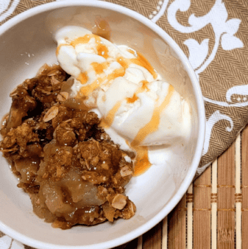 grandmas old fashioned apple crisp sitting in a white bowl with vanilla ice cream drizzled with caramel sauce
