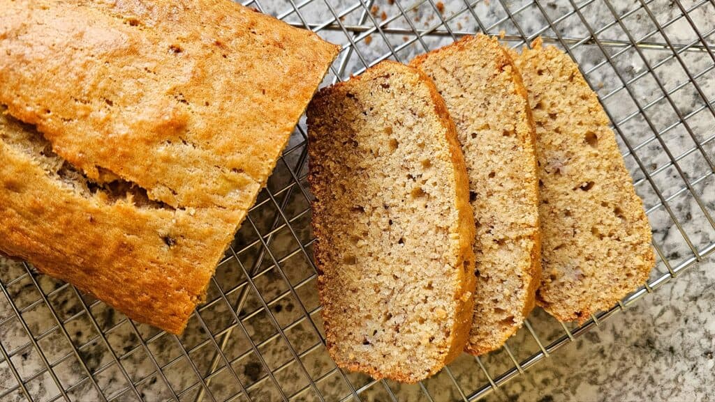 banana bread that is half sliced and half not, sitting on a cooling rack on a white and grey counter top
