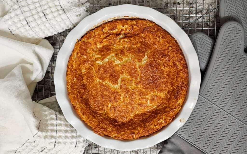 Impossible Coconut Pie Recipe (without bisquick) in a ceramic pie dish on a cooling rack with a hand towel next to it and oven mits