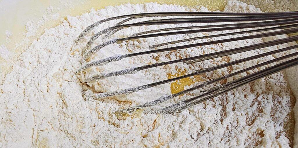 flour and baking powder on wet batter with a whisk in it