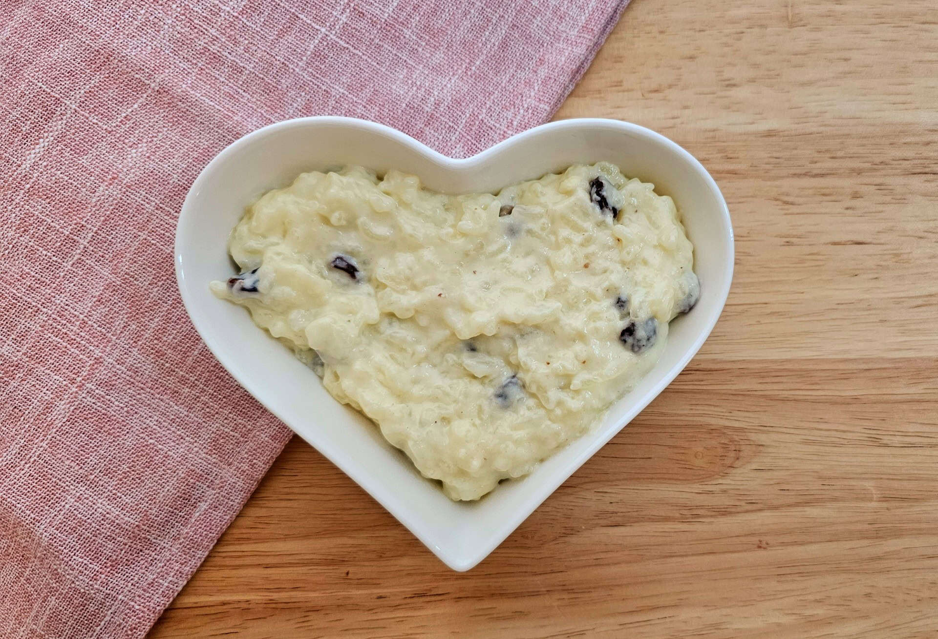 rice pudding in a white heart dish on a wooden counter top with a pink cloth napkin under half of the bowl