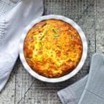 Crustless quiche in a deep pie dish on a wire rack cooling with grey oven mitts next to it