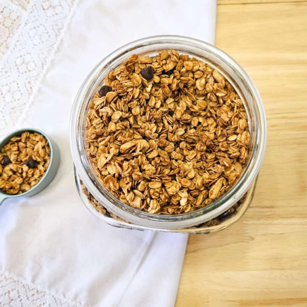 chocolate chip peanut butter granola in a large mason cookie jar on top of a wooden counter top and white cloth napkin with lace trim