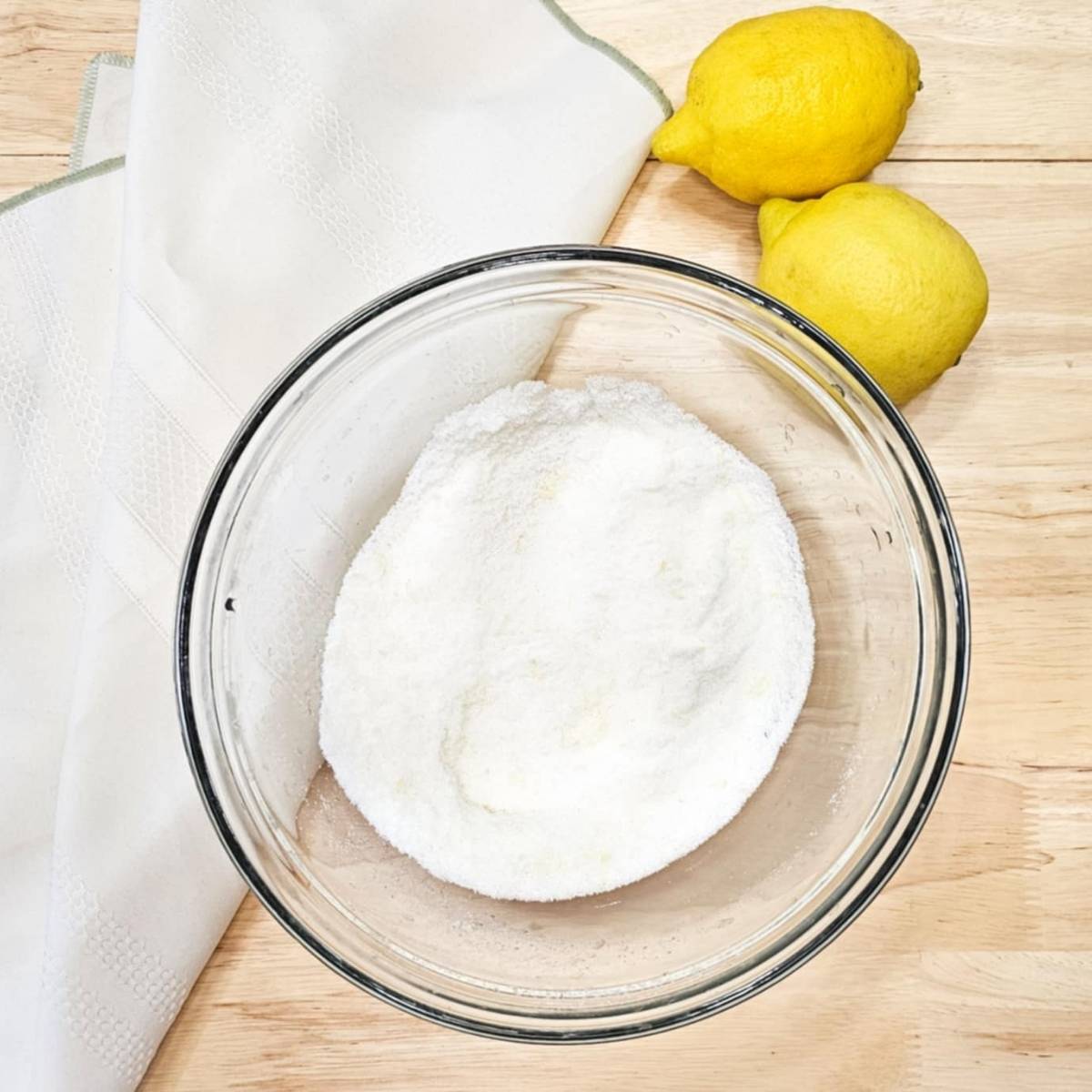 zest of a lemon pressed into sugar sitting in a glass bowl with 2 lemons next to it