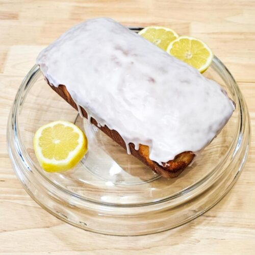 lemon loaf on a glass serving platter with sliced lemons next to it