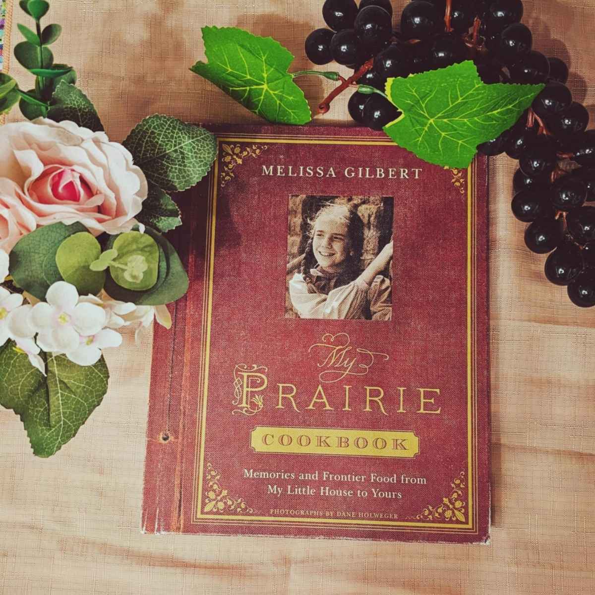 The Prairie Cook book on a pnk background with flowers and fake grapes next to it