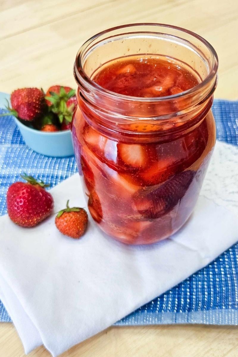 strawberry sauce in a mason jar on a blue place mat with fresh strawberries in a teal measuring cup near by on a wooden counter top