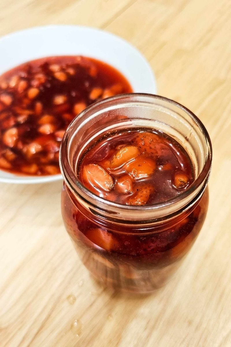 strawberry sauce in a mason jar with the leftovers in a white glass bowl behind it on a wooden counter top