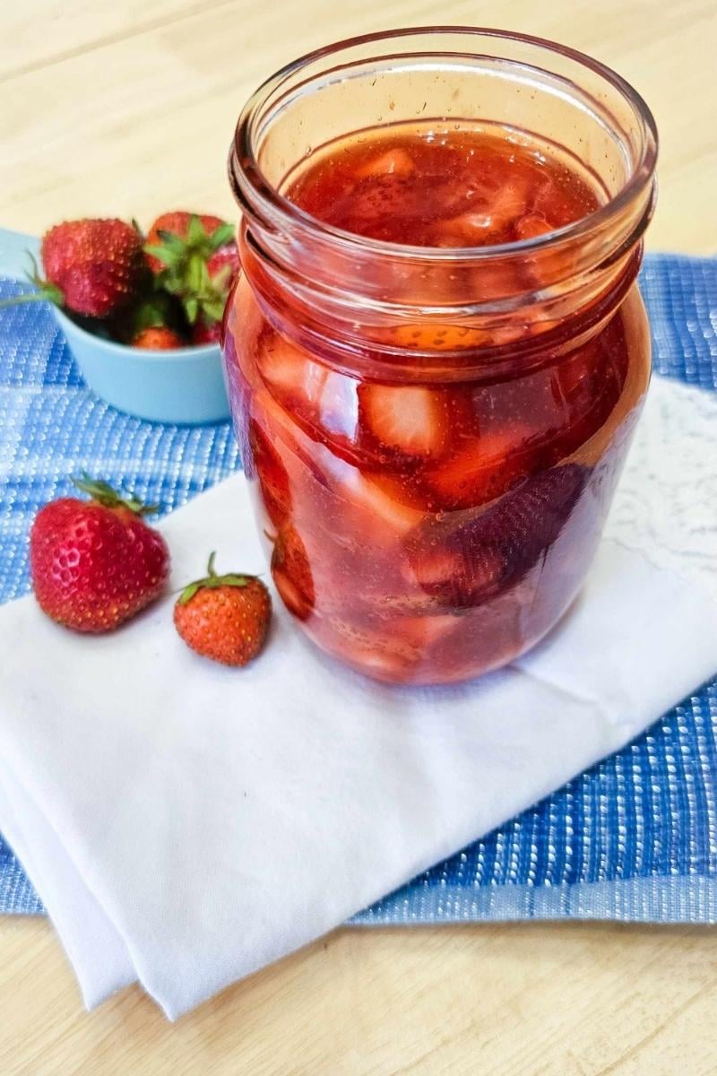 Classic strawberry sauce in a mason jar on a blue place mat and white cloth napkin with a couple of fresh strawberries next to it