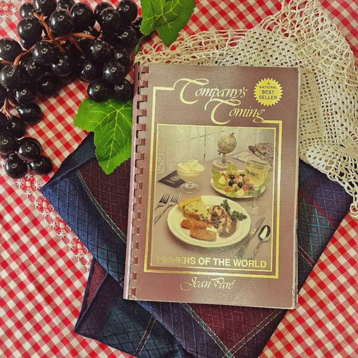 companys coming dinners of the world cookbook on a red and white table cloth with plastic black grapes next to it and a cream doily

