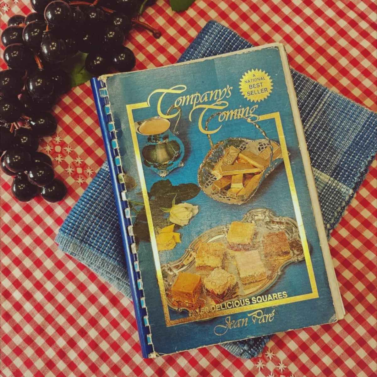 companys coming cookbook 150 delicious squares sitting on a table cloth with black fake grapes next to it
