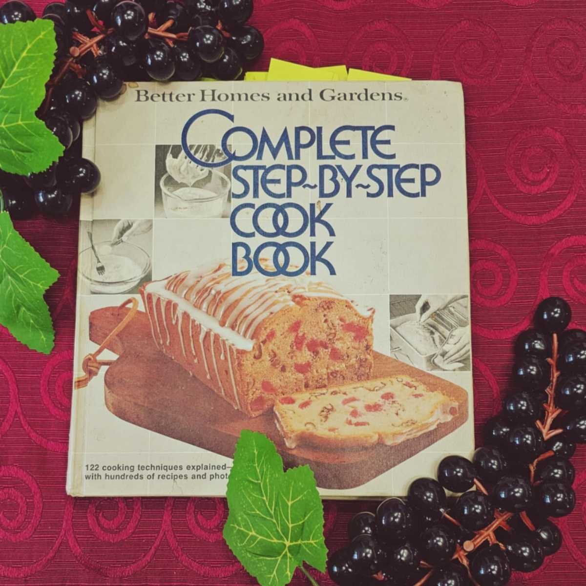 complete step by step cook book better homes and gardens cookbook on a red table cloth with plastic black grapes next to it