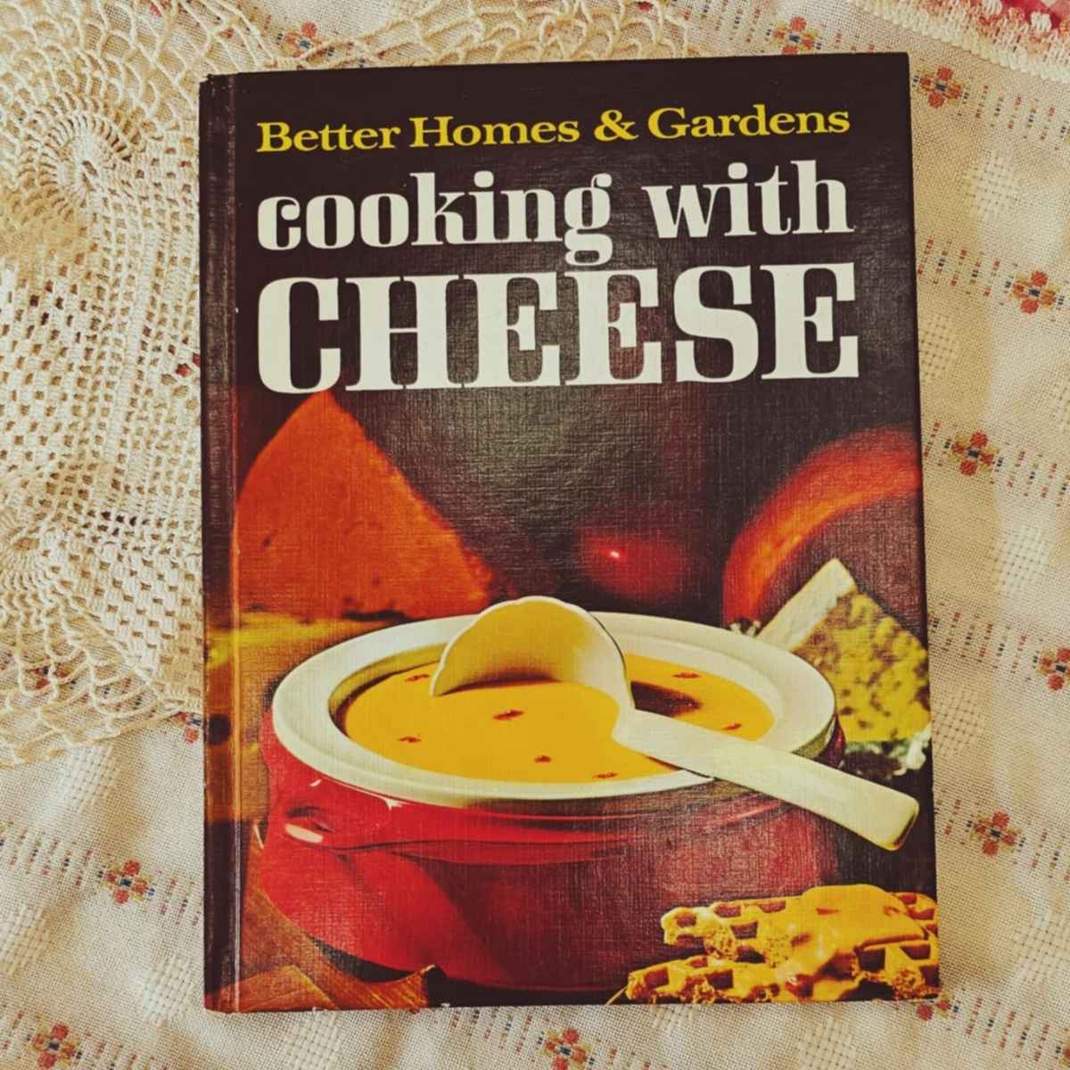 cooking with cheese better homes and gardens cookbook on a vintage table cloth with a doily to the side