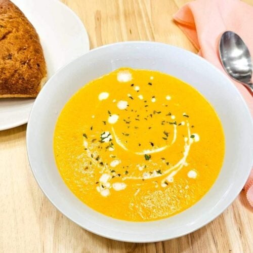 creamy carrot soup in a white bowl drizzled with cream and garnished with parsley with a spoon on a pink cloth napkin next to it and a buttered dinner roll