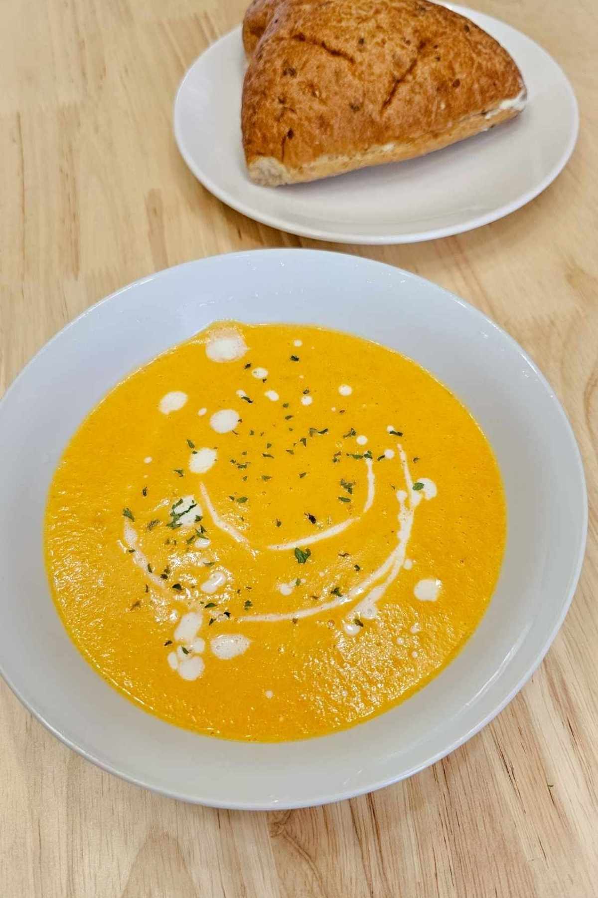 Creamy carrot soup in a white bowl, drizzled with cream and sprinkled with parsley with a buttered dinner roll near it