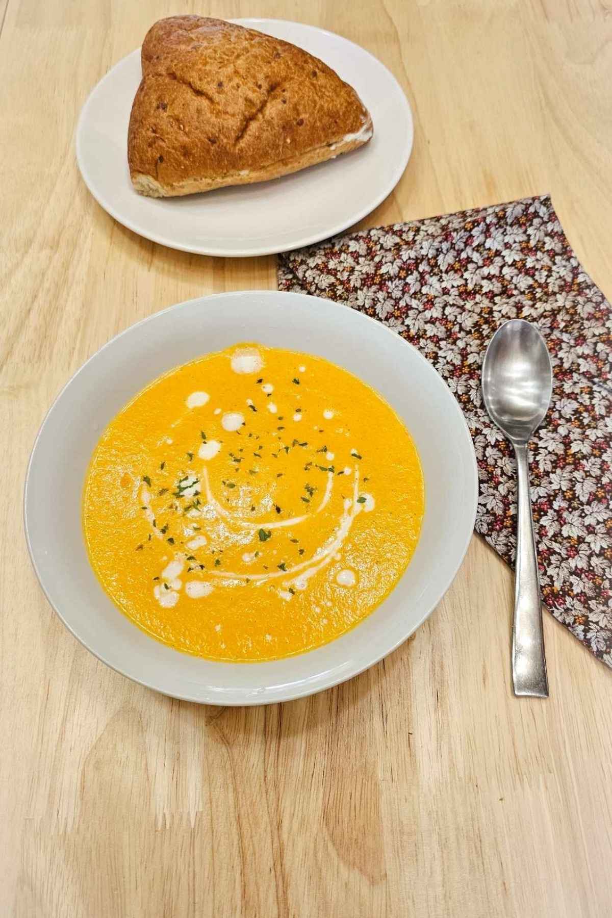 creamy carrot soup in a white glass bowl with a spoon next to it and a dinner roll