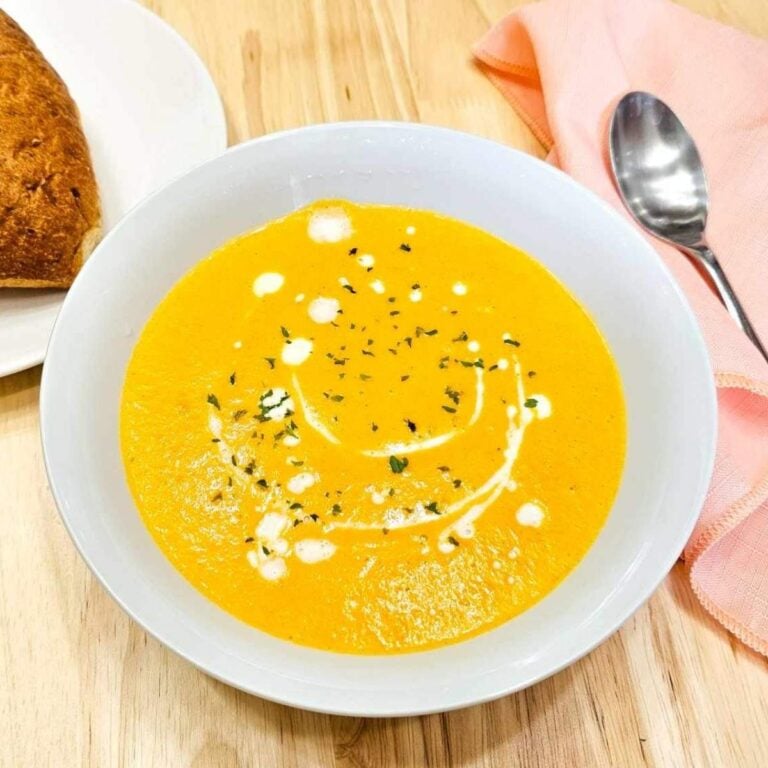 Creamy carrot soup in a white glass bowl with a pink cloth napkin that has a spoon on it with a dinner roll to the side on a plate