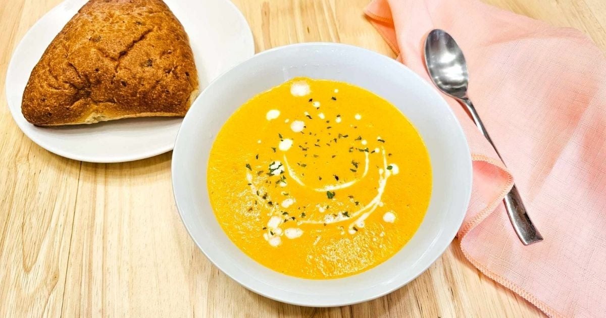 creamy carrot soup in a bowl with a pink cloth napkin next to it with a spoon and a bun on the side