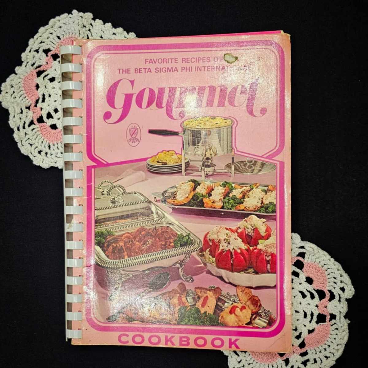 gourmet cookbook beta sigma cookbook on a black table cloth with doiles on the side