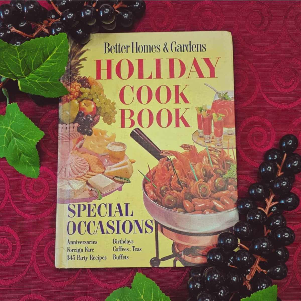 better homes and gardens cook book with a red tablecloth in the back ground and black plastic grapes with green leaves