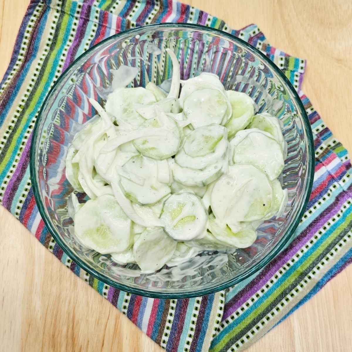 creamy cucumber salad in a clear glass bowl on a colourful table mat on a wooden counter