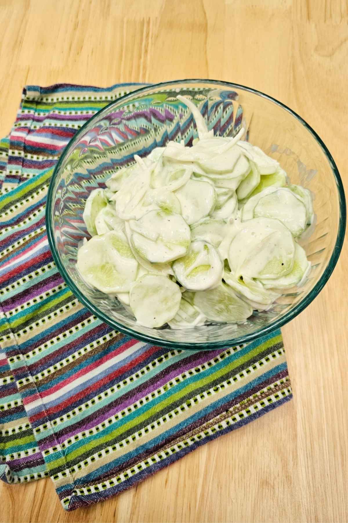sour cream cucumber salad in a clear glass bowl on a colourful place mat on a wooden table