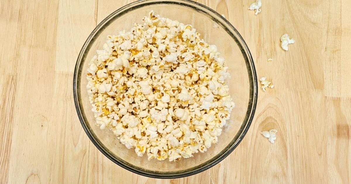 stove top popcorn in a clear glass bowl on a wooden counter top with some popped kernels to the side of it