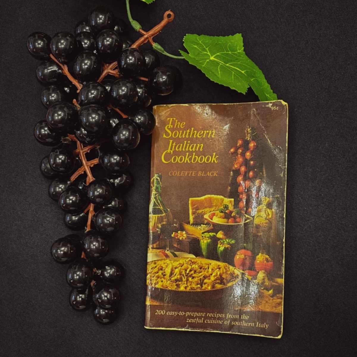 The southern italian cook book by colette black on a black table cloth with black fake grapes next to it with green leaves