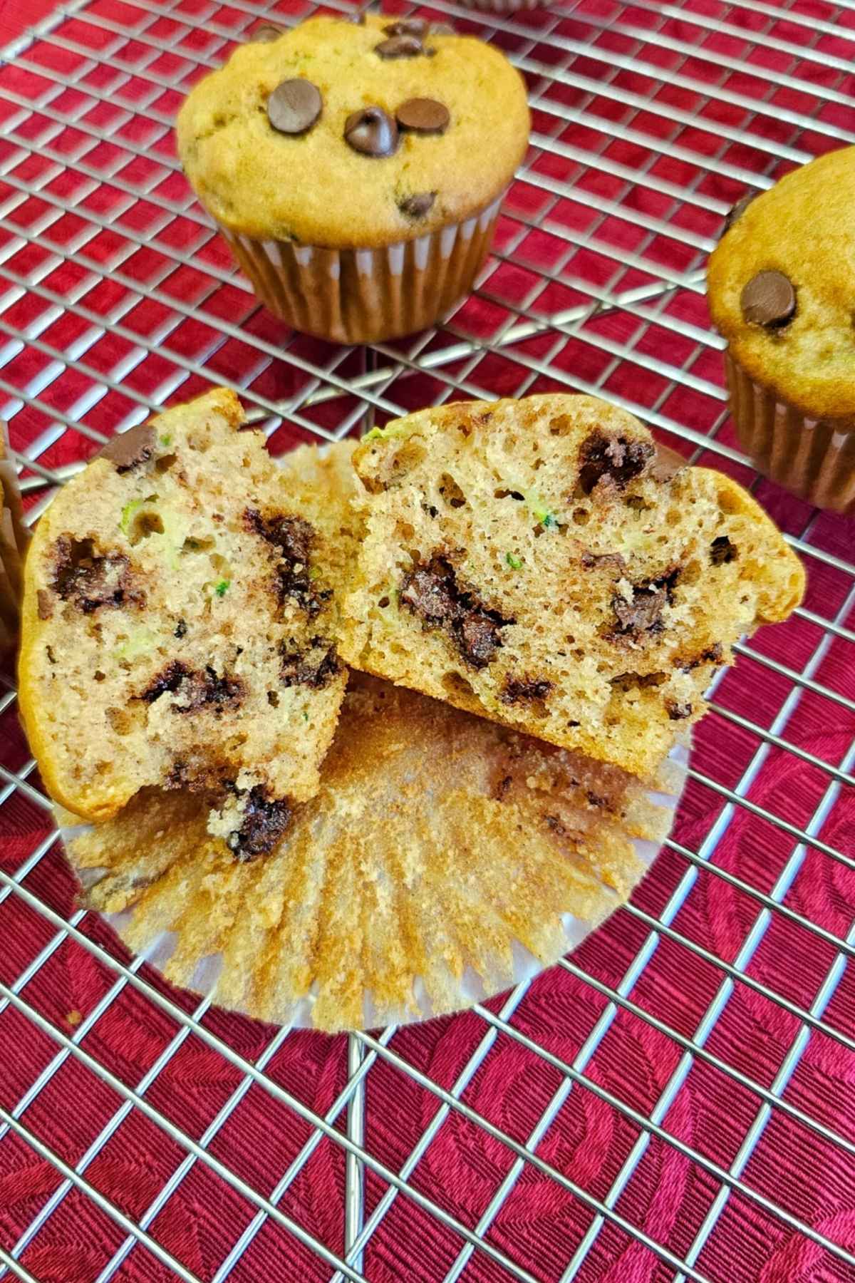 chocolate chip zucchini muffin sliced in half on a wire rack sitting on a muffin liner with a red table cloth under the wire rack