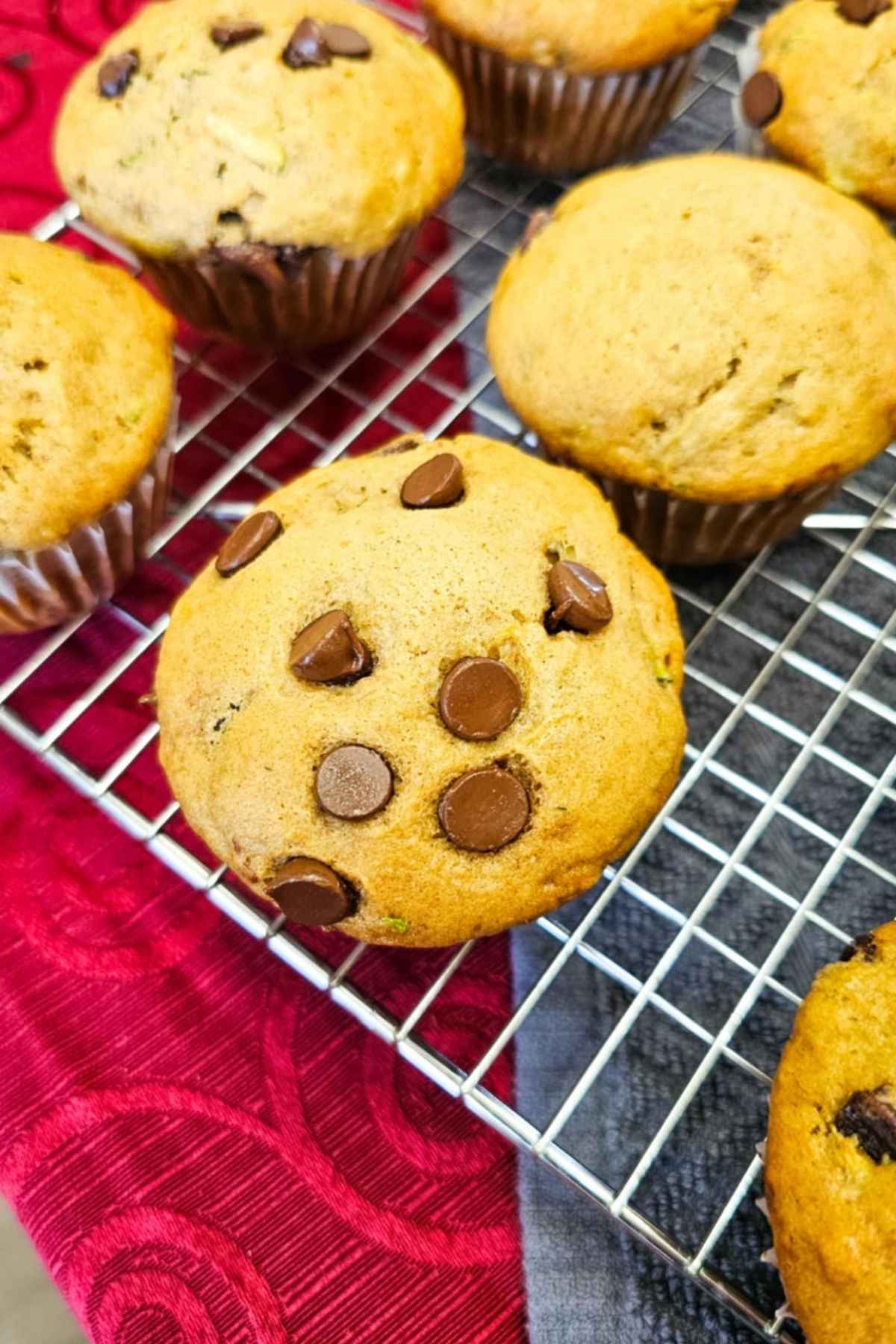 7 chocolate chip zucchini muffins on a wire rack topped with chocolate chips and on a red vintage table cloth