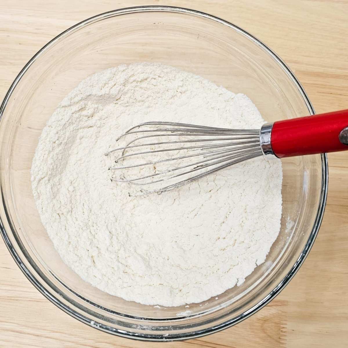 flour, baking powder, baking soda, salt and ginger in a glass clear bowl on a wooden counter top with a whisk in the bowl